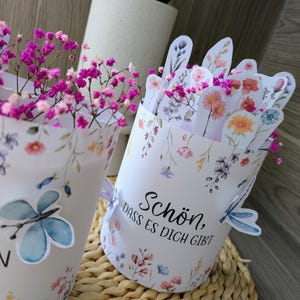 May include: Two white paper containers with floral and butterfly designs, filled with pink flowers. One container has the text "Sch&ouml;n, dass es dich gibt". The containers are on a woven surface.