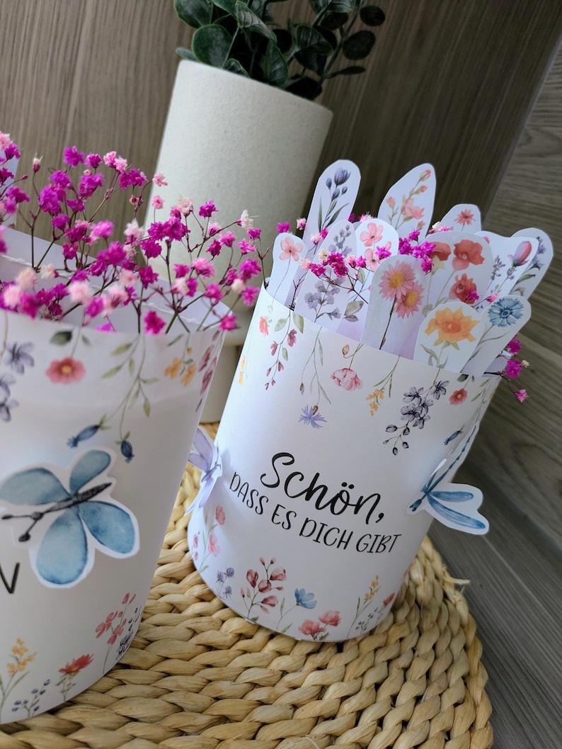 May include: Two white paper containers with floral and butterfly designs, filled with pink flowers. One container has the text "Sch&ouml;n, dass es dich gibt". The containers are on a woven surface.