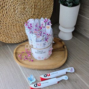 May include: A floral-themed birthday gift set. The set includes a round container with a floral design and the words "Happy Birthday". Two white and red candy bars are also included, along with a blue butterfly decoration.