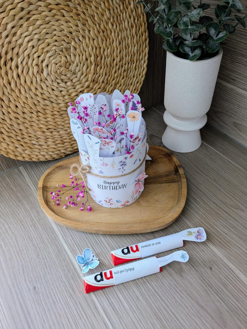 May include: A floral-themed birthday gift set. The set includes a round container with a floral design and the words "Happy Birthday". Two white and red candy bars are also included, along with a blue butterfly decoration.
