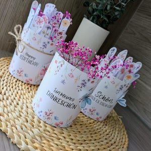 May include: Three white floral-patterned paper containers with pink flowers. One container has "Happy Birthday" text, another says "Ein kleines Dankesch&ouml;n", and the third says "Sch&ouml;n, dass es dich gibt". The containers are on a woven mat.