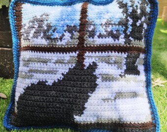 May include: A crocheted pillow with a blue border and a pixelated design of a winter scene. The design features a snowy landscape with trees and a dark silhouette of a figure.
