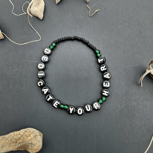 May include: Black beaded bracelet with green beads and white letters spelling "dedicate your heart".