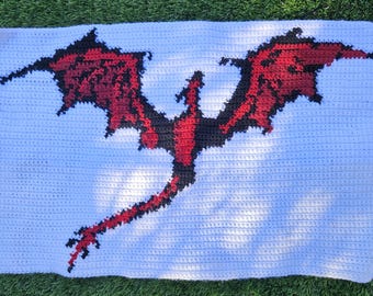 May include: A white crocheted blanket with a red and black dragon design. The dragon is flying with its wings spread out.