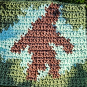 May include: Crocheted square with a brown silhouette of a Bigfoot on a light blue background. The square is surrounded by a green border.