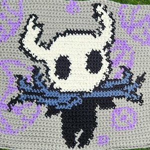 Può includere: Quadrato all'uncinetto con un design di personaggio con testa bianca, occhi neri e sciarpa blu. Il personaggio è ambientato su uno sfondo grigio con accenti viola. Il lavoro all'uncinetto è dettagliato e strutturato.