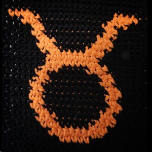 May include: A close-up of a black crocheted item with an orange Taurus zodiac symbol. The symbol is the focal point, set against the dark background, highlighting the detailed stitch work and contrasting colours.