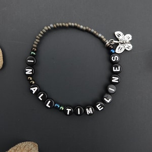 May include: A black beaded bracelet with white letter beads spelling "IN ALL TIMELESS". The bracelet has a silver butterfly charm and a dark gray beaded band. The bracelet is on a dark gray surface.