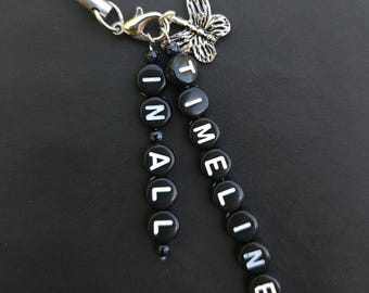 May include: A black beaded charm with the words "IN ALL TIMELINES" in white letters. It has a silver butterfly charm and a silver clasp. The charm is attached to a black cord.