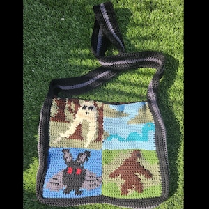 May include: A crocheted shoulder bag with a black and gray strap. The bag is made of squares with different colored crocheted designs, including a brown creature, a blue creature, a brown creature in a tree, and a blue creature with red eyes.