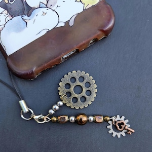 May include: A brown phone case with a cat illustration, a bronze gear, and a charm with beads and a key-shaped pendant. The charm is attached to a black cord. The gear and charm have a steampunk aesthetic.