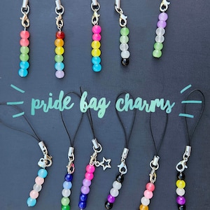 May include: A collection of colorful beaded bag charms, each with a different rainbow or pride flag color scheme. The charms are attached to black cords with silver-colored clasps. The text "pride bag charms" is written in a teal script.