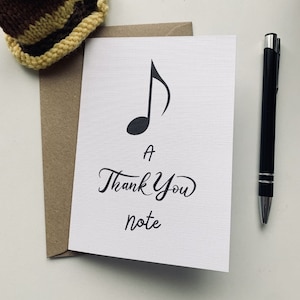 May include: A white card with a black musical note and the text "a thank you note". The card is sitting on a brown envelope. A brown and yellow knitted hat and a black pen are also in the image.