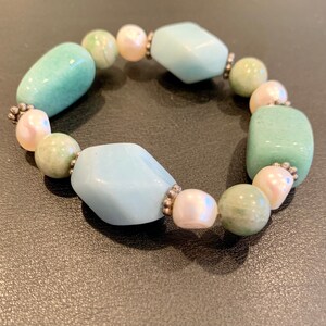 May include: A beaded bracelet featuring a mix of light blue, green, and off-white stones. The bracelet includes rounded and oblong beads, with silver-colored spacers between the stones. The bracelet is designed to be worn around the wrist.