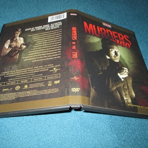 May include: DVD case for the film "Murders in the Zoo." The cover features a man in a suit and the title in red. The back of the case has text and an image of a person holding a weapon. The case is black and gold.
