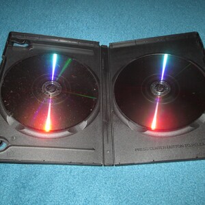 May include: An open black DVD case with two reflective discs inside. The discs have a rainbow-like sheen, with red, orange, green, and blue light reflecting off their surfaces. The case is on a blue surface.