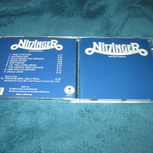 May include: A blue and white CD case with the title "Nitzinger" and the text "One Foot in History". The back of the case lists the songs on the album, including "Take a Picture", "Motherlode", "Good Bless the Pervert", "Earth Eater", "Driftwood", "Let the Living Grow", "The Cripple Gnat Bounce", "One Foot in History", "Uncle John", and "Texas Blues/Jelly Roll".