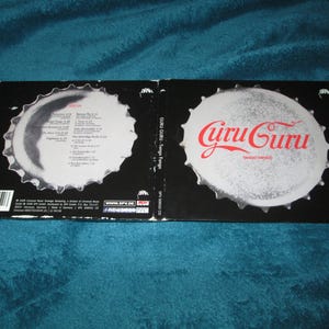 May include: A CD case with a black background and a white bottle cap design. The front of the case features the text "Guru Guru" in red letters. The back of the case has a list of songs and credits.
