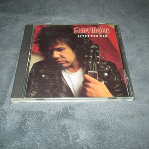 May include: A compact disc case with a red, white, and blue background. The cover features a photo of Gary Moore, a musician, holding a guitar. The title of the album is "Gary Moore After the War".