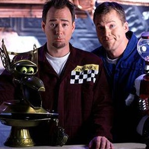 May include: Two men stand in front of a silver and blue background. The man on the left is holding a gold trophy with a robot head. The man on the right is holding a red robot with a clear glass head. The robot is holding a red and white sign that says "9".
