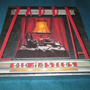 May include: Zapp "The Old Masters Box One" album cover. The album art features a still life with a lamp, shoes, and a purse on a table in front of a red curtain. The album title and band name are displayed in red and silver.
