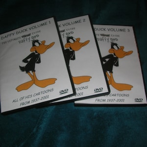 May include: Three DVD cases featuring Daffy Duck cartoons. The cases are black with white text and images of Daffy Duck. The text on the cases reads "Daffy Duck Volume 1", "Daffy Duck Volume 2", and "Daffy Duck Volume 3". The text also reads "The Ultimate Guide Daffy DVD" and "All of His Cartoons From 1937-2001".