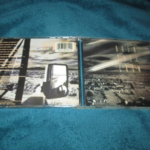 May include: A CD case with a black and white photo of a desert landscape. The front cover has the text "Rain In The  /  Dry Season" and the back cover has a list of songs.