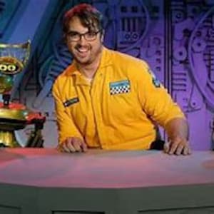 May include: A man in a yellow jumpsuit stands behind a counter with two robots. The robot on the left is gold and has a round head with a single eye. The robot on the right is red and has a round head with a single eye. The man is smiling and looking at the camera.