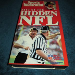 May include: A VHS tape with a red and white label featuring a photo of a football coach and referee. The title is "Best of the Hidden NFL" with the Sports Illustrated logo. The tape is distributed by NFL Films Video.