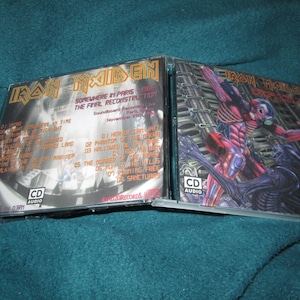 May include: A double CD set of Iron Maiden's "Somewhere in Paris: The Final Reconstruction" concert. The front cover features a black and white photo of the band performing on stage. The back cover features a colorful illustration of a red and black creature with a metal helmet.