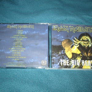 May include: A CD case of the Iron Maiden album "The Big Heat" is open, revealing the CD and its cover art. The cover features a monster with yellow skin and a cityscape background. The tracklist is visible on the back.