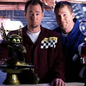 May include: Two men stand in front of a silver and blue background. The man on the left is holding a gold trophy with a robot head. The man on the right is holding a red robot with a clear glass head. The robot is holding a red and white sign that says "9".