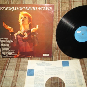 May include: A vinyl record album cover featuring David Bowie. The cover is brown with a picture of Bowie in a sparkly outfit. The album title "The World of David Bowie" is printed in white letters at the top of the cover. The album is by Decca Records.