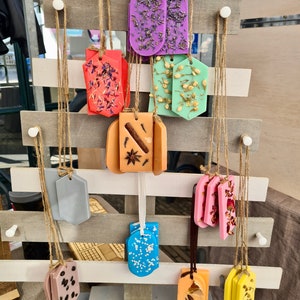 May include: A display of various scented wax melts, each with a different design and color. The wax melts are hanging from a wooden rack with twine. The wax melts are shaped like tags and have different floral and spice designs.