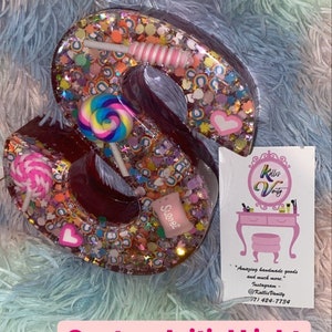 May include: A decorative letter "S" filled with colorful confetti, candies, and lollipops. The letter is a custom initial light with the text "Custom Initial Light @kallisvanity" on a pink banner. A business card is also visible.