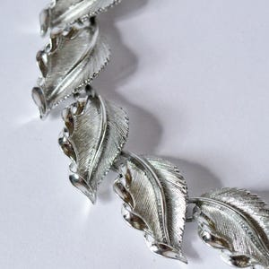 Könnte beinhalten: Eine silberfarbene Metallhalskette, die aus einer Reihe von miteinander verbundenen blattförmigen Gliedern besteht. Jedes Glied weist eine texturierte, gebürstete Oberfläche mit polierten, glatten Kanten auf, die ein sich wiederholendes botanisches Muster bilden.