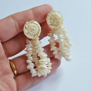 Könnte beinhalten: Ohrringe aus cremefarbenen Muscheln. Jeder Ohrring hat einen kreisförmigen Stecker mit einem Spiralmuster, von dem eine Kaskade aus kleinen Muscheln hängt. Die Ohrringe haben eine helle, natürliche Farbe und das Design ist filigran und zart.