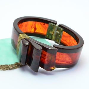 Könnte beinhalten: Ein aufklappbarer Armreif mit einem Design aus dunkelbraunem und durchscheinendem orangefarbenem marmoriertem Harz. Der Armreif verfügt über ein dekoratives goldfarbenes Metallakzent mit gerippten Details und einen funktionalen Metallscharniermechanismus an der Seite.