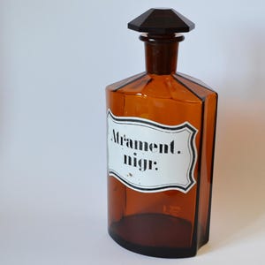 Könnte beinhalten: Eine bernsteinfarbene Glasflasche mit einem dunkelbraunen Verschluss. Die Flasche hat eine rechteckige Form und ein weißes Etikett mit der Aufschrift "Atrament. nigr:". Ein Vintage-Sammlerstück.