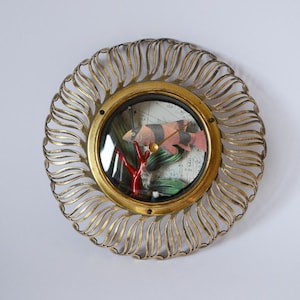 Op de afbeelding: Een ronde, goudkleurige wandklok met een decoratief, zonvormig frame. De wijzerplaat heeft een miniatuur diorama met rode koraal, vissen en een kaartachtergrond. De klok is aan een witte muur bevestigd.