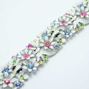 Könnte beinhalten: Ein Gliederarmband aus silberfarbenem Metall mit weißen Emaille-Blumenmotiven. Jede Blume ist mit einem funkelnden Strassstein in der Mitte in den Farben Pink, Hellblau oder Limettengrün verziert, was ein florales Muster ergibt.
