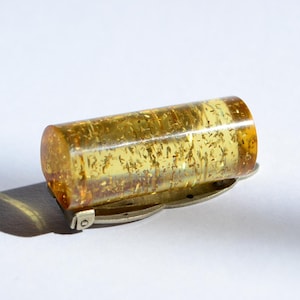 Könnte beinhalten: Eine zylindrische Brosche mit einem durchscheinenden, goldgelben Bernstein, der sichtbare Einschlüsse aufweist. Der Bernstein ist auf einer silberfarbenen Metallnadel mit Scharnierverschluss montiert und zeigt eine polierte Oberfläche.