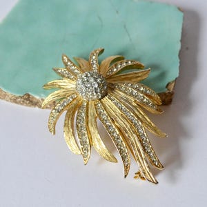 Könnte beinhalten: Goldfarbene Brosche in floralem Design, mit einem zentralen Cluster aus klaren Strasssteinen und strahlenförmigen Blütenblättern, die mit kleineren Strasssteinen verziert sind. Die Brosche ist vor einem hellblauen und weißen Hintergrund platziert.