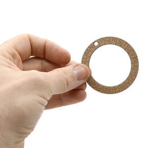 May include: A wooden cipher wheel with a circular design and an alphabet around the edge. The wheel has a small hole at the top for hanging. The wheel is held in a hand against a white background.