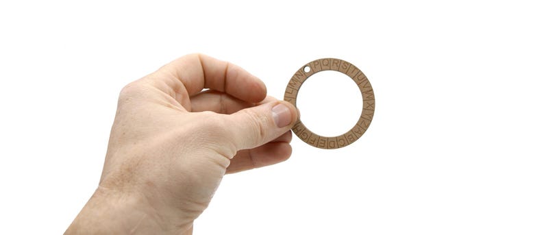 May include: A wooden cipher wheel with a circular design and an alphabet around the edge. The wheel has a small hole at the top for hanging. The wheel is held in a hand against a white background.