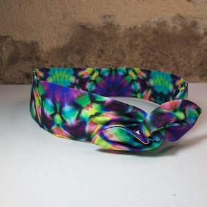 Peut inclure: Un bandeau tie-dye coloré avec un nœud sur un côté. Le bandeau est en tissu et présente un motif psychédélique dans des tons de bleu, vert, violet et jaune.