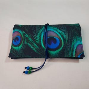 Puede incluir: Estuche de tela enrollado con un diseño de plumas de pavo real en tonos verdes, azules y morados. Se asegura con un cordón azul marino y adornado con cuentas azules y verdes. La tela tiene un fondo oscuro.
