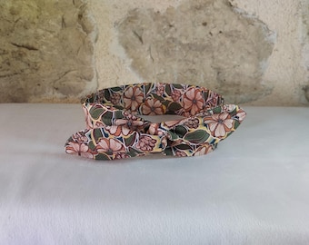 May include: A floral patterned headband with a knotted bow. The fabric features a repeating pattern of pink and peach flowers with green leaves on a yellow and dark green background. The headband is on a white surface.