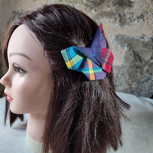 May include: A colorful plaid hair bow with a purple, teal, yellow, and red plaid pattern. The bow is tied in a knot and has a simple design.