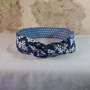 May include: A navy blue headband with a floral pattern of white flowers and red accents. The interior lining features a blue and white geometric design. This knotted headband is a stylish hair accessory.